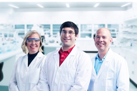 2 men and 1 woman posing in a lab wearing lab coats and glasses