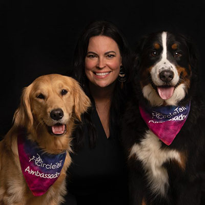 A woman with black hair is posing next to two dogs wearing a CircleTail Ambassador handkerchief